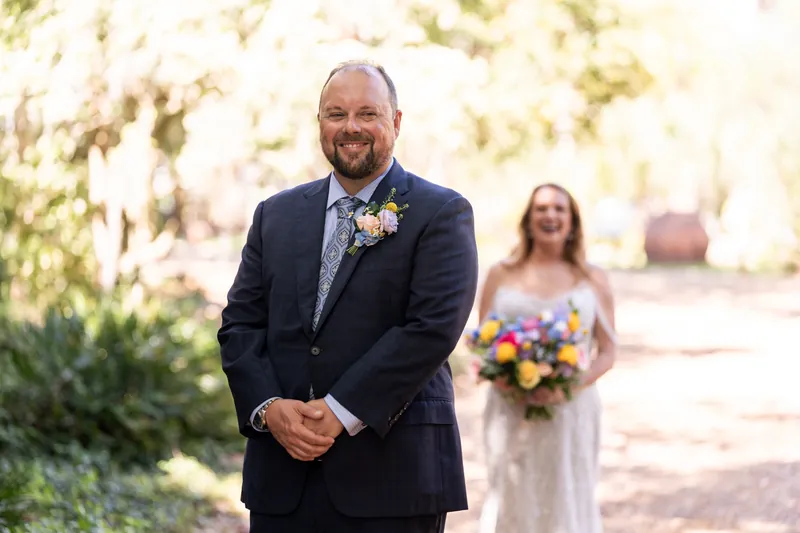 Von smiles at the camera wearing a navy suit with a boutonniere on a tree-lined path.