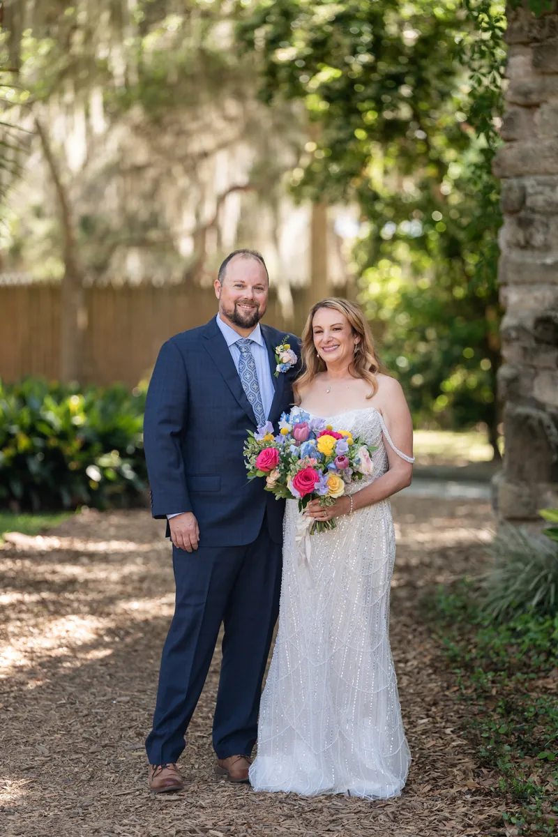 Von and Danielle stand together on a gravel path with Danielle holding a colorful bouquet of roses and wildflowers.