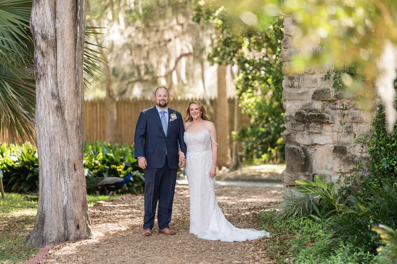 Von in a navy suit and Danielle in a white wedding dress walk hand-in-hand down a tree-lined path.