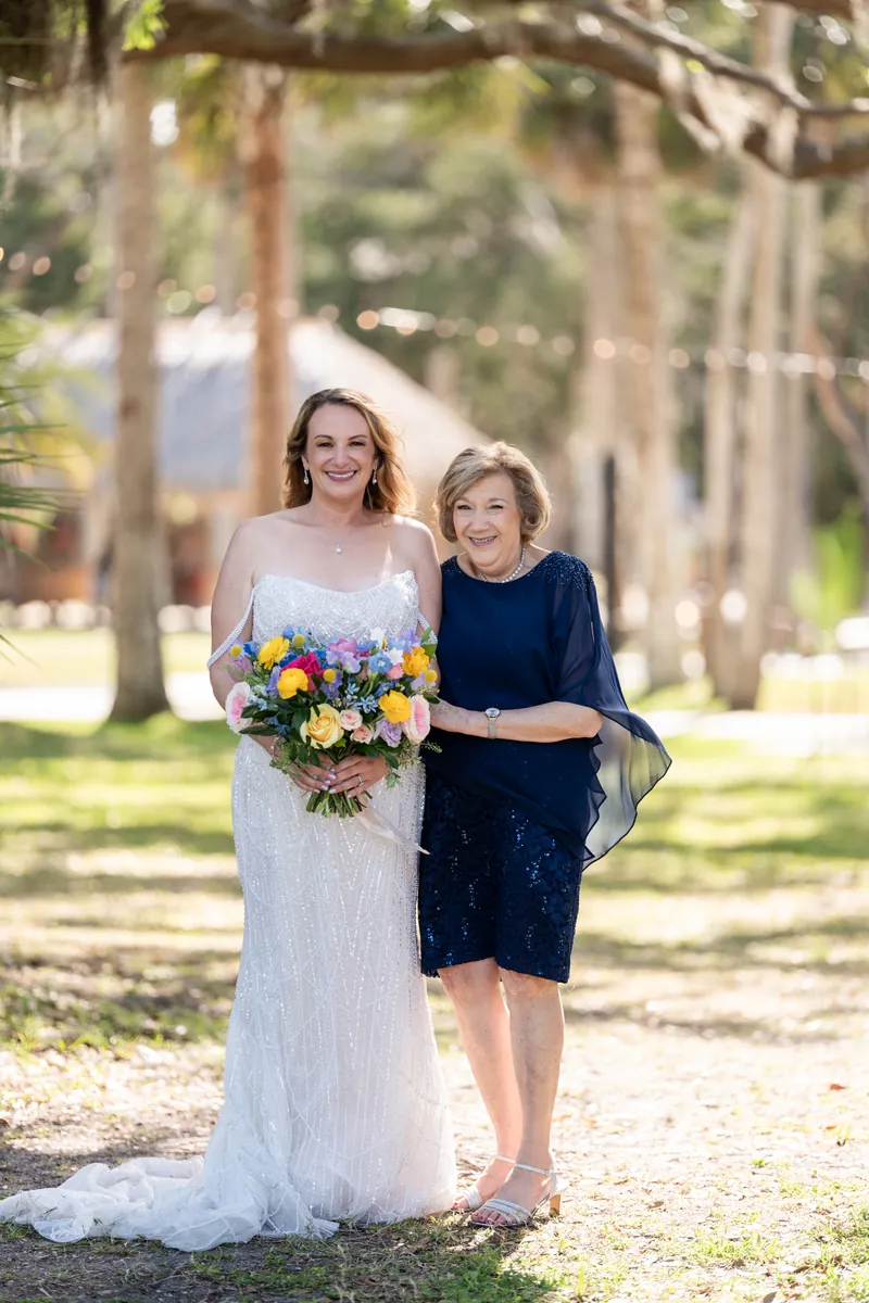 Danielle holds a colorful bouquet while posing with an older woman in a navy dress beneath tall trees.