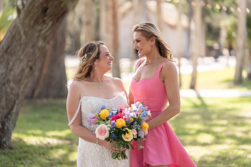 Danielle in her white gown and a woman in a pink dress hold a vibrant mixed-flower bouquet while talking outdoors.