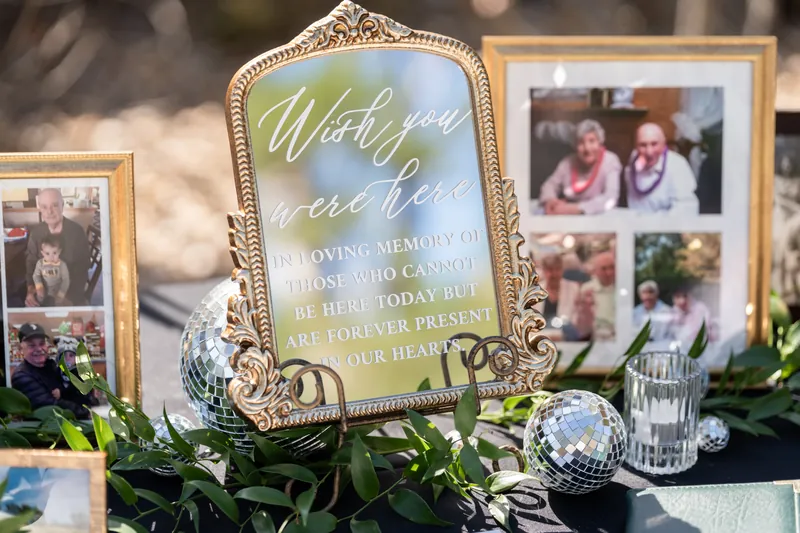Framed family photos and a mirrored "Wish you were here" sign arranged on a table with flowers and glassware.