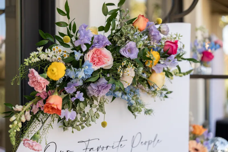 Lush floral arrangement with pink, purple, yellow, and coral roses on white sign reading "Our Favorite People.