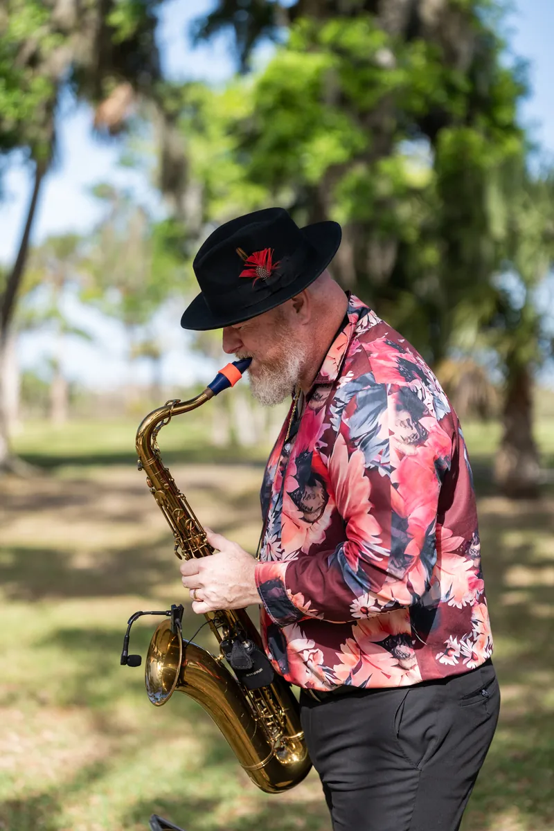 Saxophonist in pink floral shirt and black hat performs outdoors under palm trees.