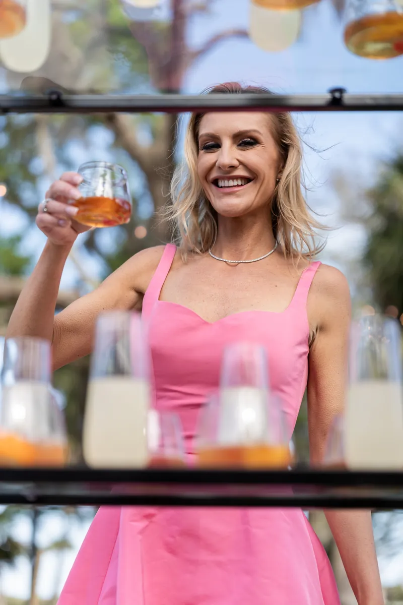 Woman in pink sleeveless dress holds champagne flute at outdoor reception with blurred background.