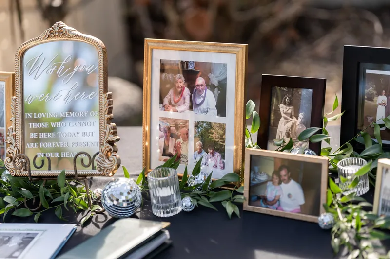 Close-up of memorial table with ornate gold-framed mirror, family photographs in various frames, and greenery.