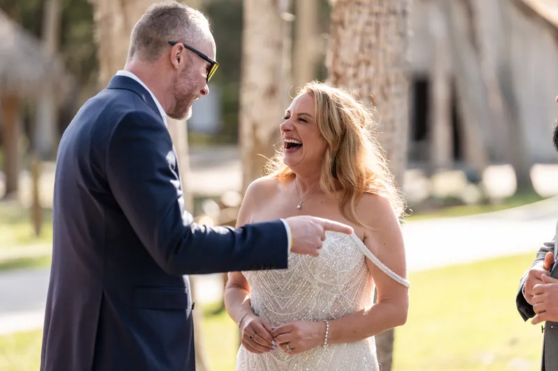 Von in a navy suit and Danielle in a white lace dress laugh together during their outdoor ceremony.
