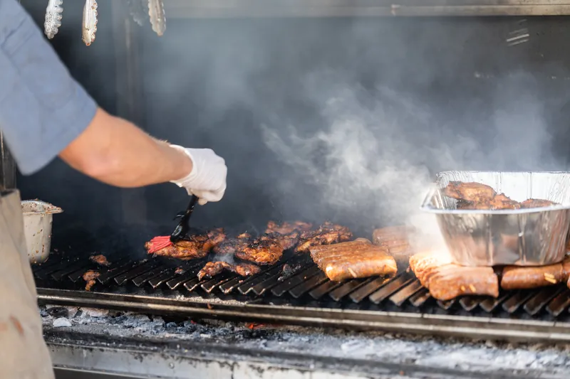 Someone in a white dress shirt tends to grilled meats smoking on a barbecue.