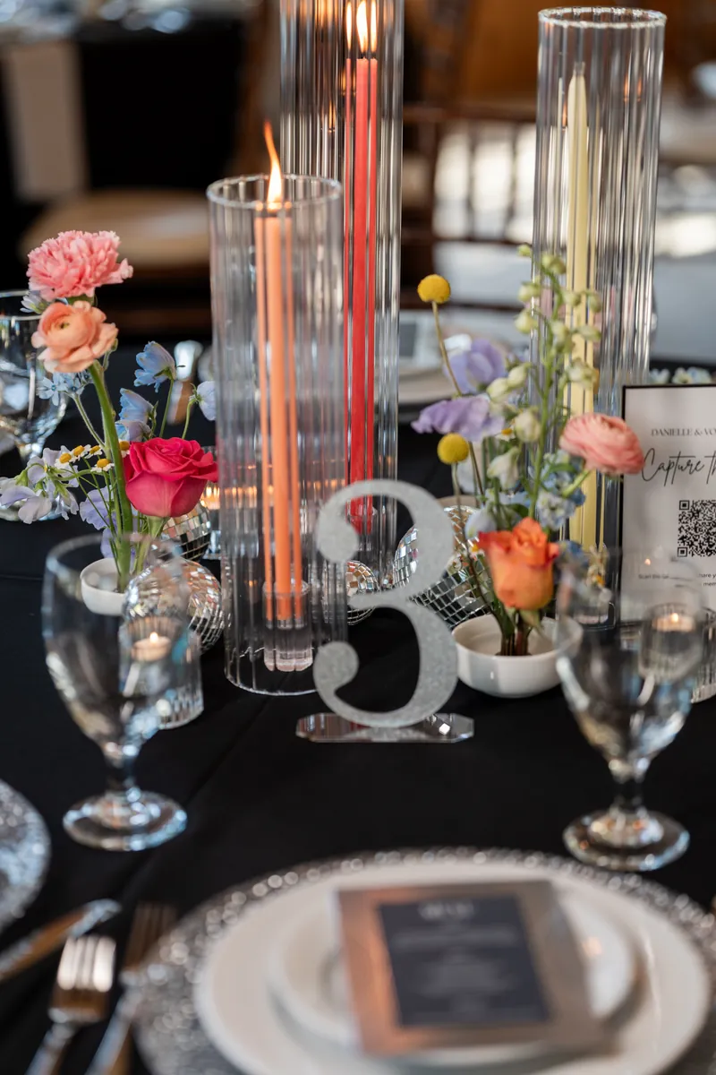 Tall glass vases with pink and red flowers and copper-colored candles on a black table.