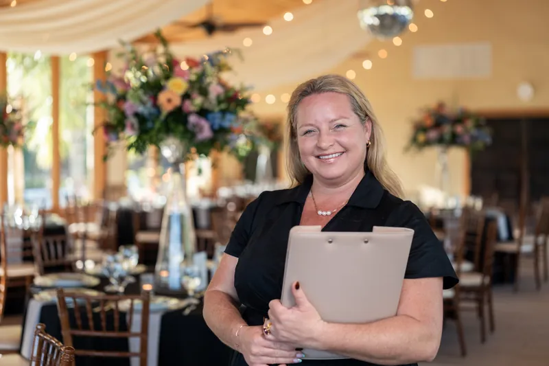Woman in black holding a tablet smiles in a decorated wedding reception hall with hanging florals.