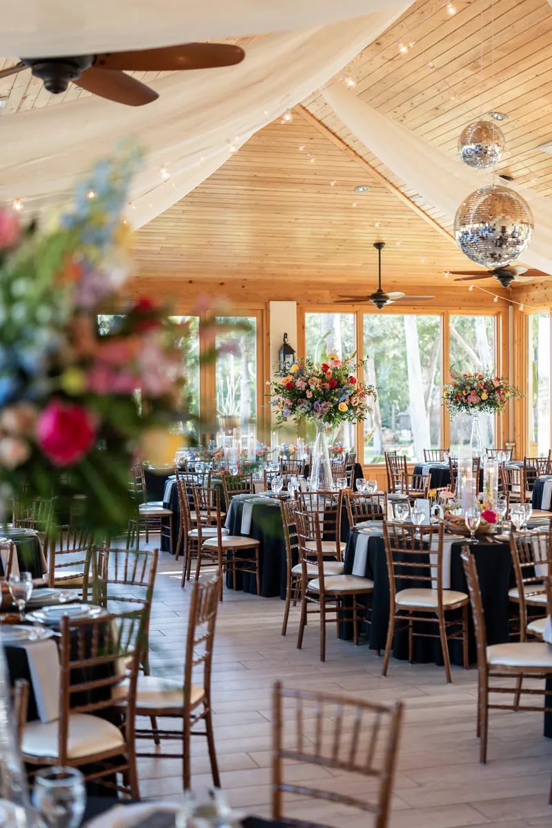 Wooden reception hall with draped fabric ceiling, chiavari chairs at round tables set with black linens and floral centerpieces.