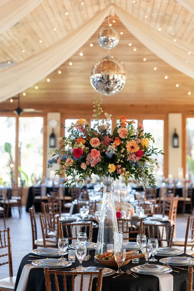 Close-up of black-linened table with white place settings, tall floral centerpiece in coral and yellow, and hanging crystal chandeliers above.