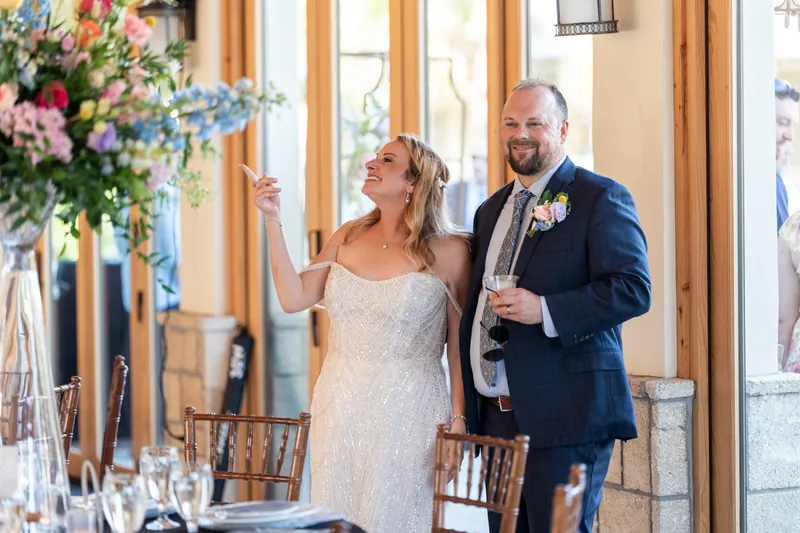 Danielle in beaded wedding dress and Von in navy suit with boutonniere stand together in front of tall floral arrangement.