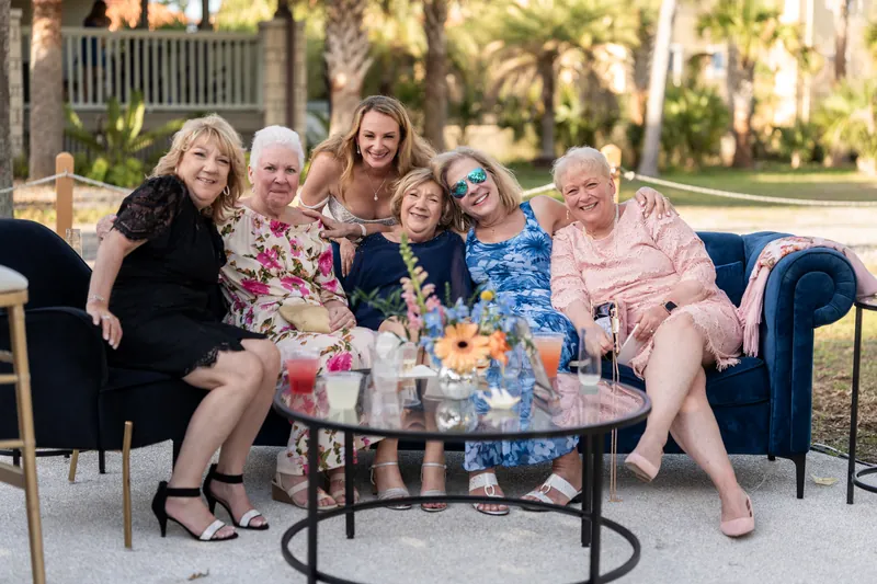 Six women sit together on a blue couch with a floral arrangement on the table in front of them under palm trees.