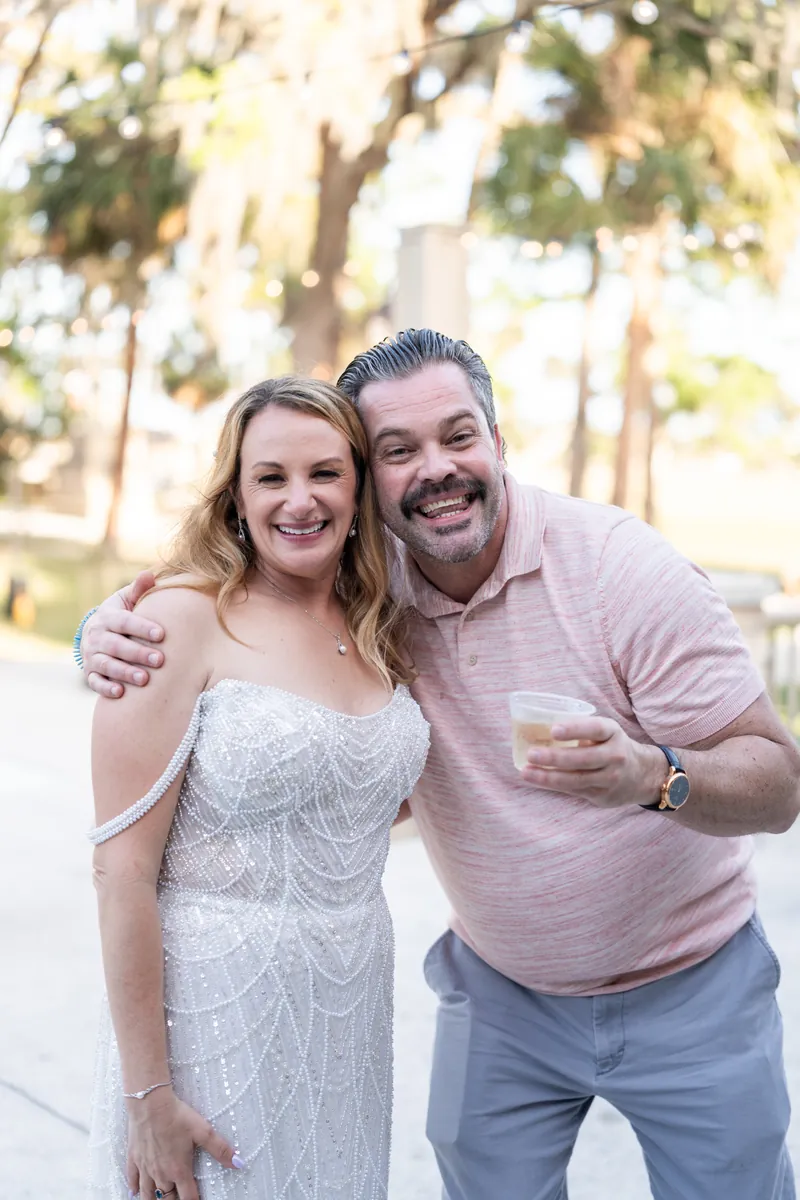 Danielle in a beaded white wedding dress smiles with a guest in a pink shirt holding a drink outdoors.