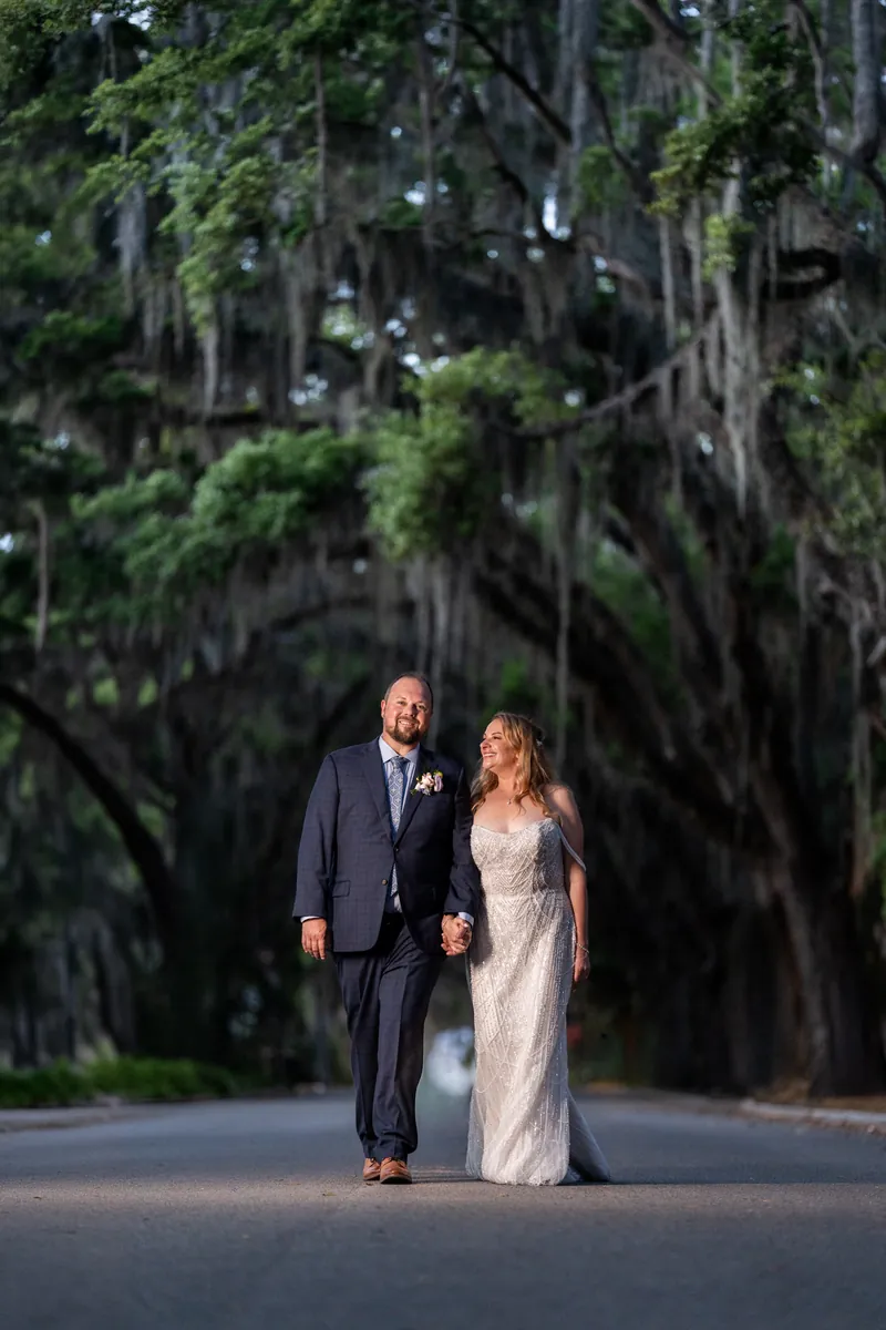Von in a dark suit and Danielle in a white wedding dress walk hand-in-hand under a canopy of Spanish moss.