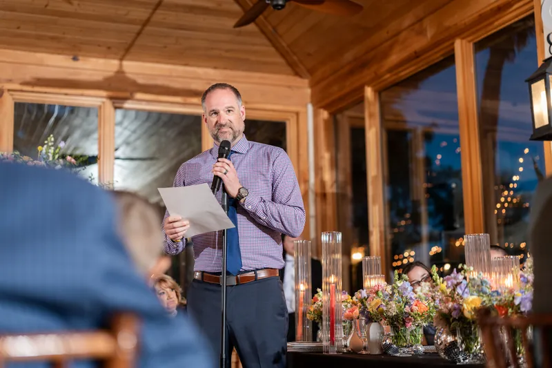 A man in a purple shirt speaks at a microphone during an indoor reception with floral centerpieces visible.