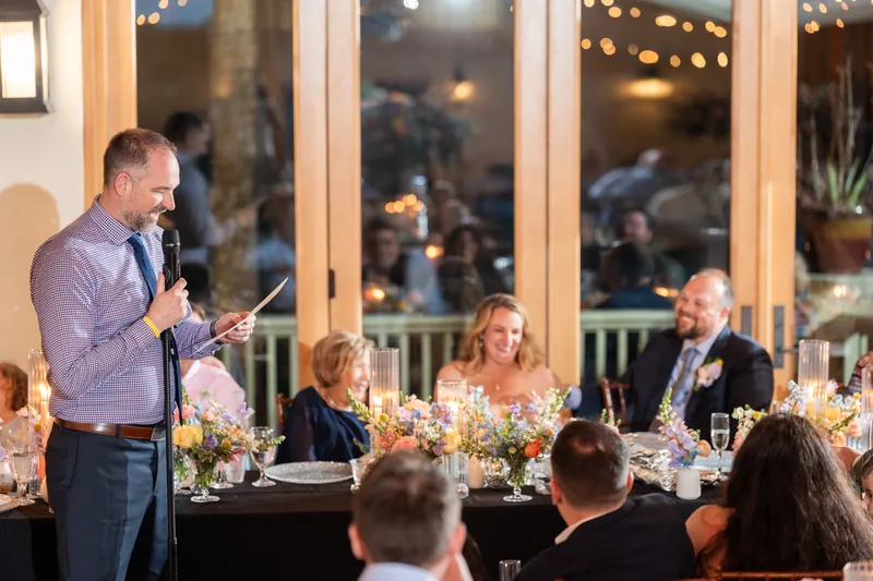 Man in lavender shirt speaking at microphone during indoor reception with guests seated at long tables.