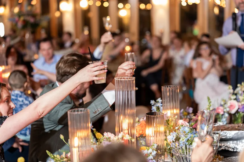 Guest raising glass for toast at candlelit dinner table with floral centerpieces and string lights.