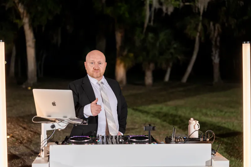 DJ in tuxedo at outdoor nighttime setup with laptop and turntables under palm trees.