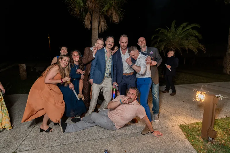 A group of wedding guests pose together outdoors at night, some crouching, with palm trees visible.