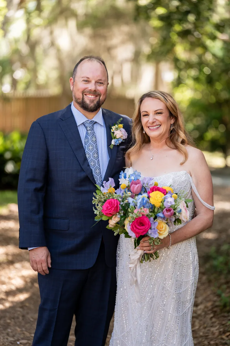 Von in a navy suit and Danielle in a beaded white dress smile while holding a vibrant bouquet under dappled tree shade.