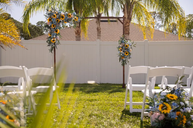 Wooden arch with hanging sunflower and dusty blue garlands frames a grass lawn set with white folding chairs beneath palm trees.