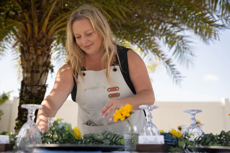 Person in a cream apron arranges yellow flowers in a vase outdoors beneath a palm tree.