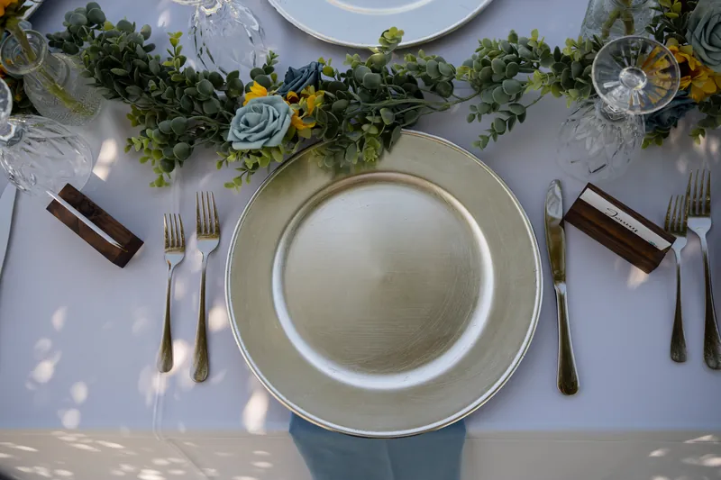 Overhead view of place settings with white plates, greenery runners, sunflower stems, and string lights on a white tablecloth.
