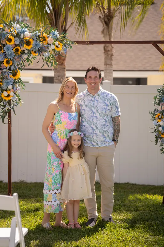 Danielle and Von stand with a child beneath a flower-decorated arch on a manicured lawn, both smiling at the camera.