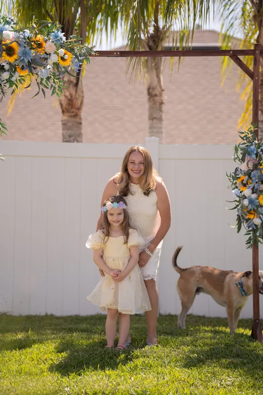 A woman and child pose together on the grass in front of the sunflower arch, with a dog walking nearby.