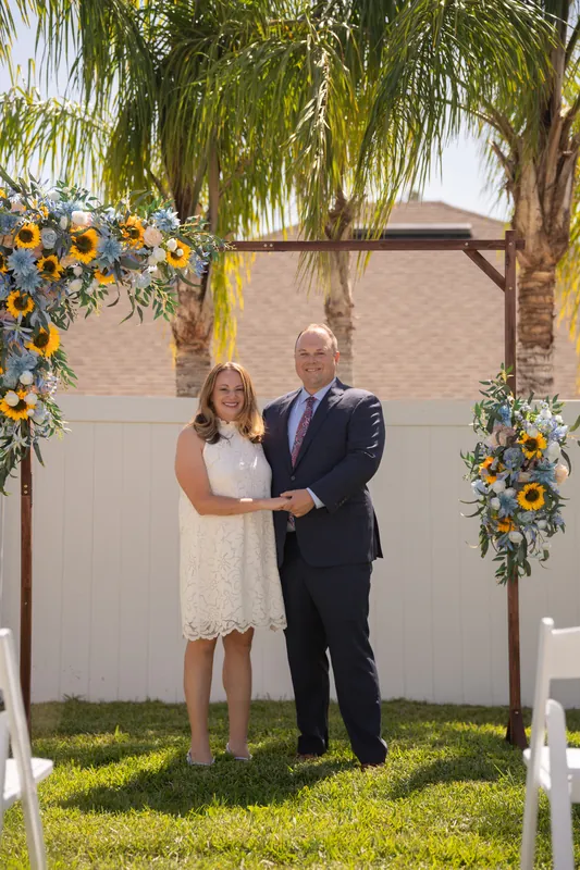 Danielle and Von hold hands beneath a wooden arch draped with sunflower garlands under palm trees at their Airbnb ceremony.