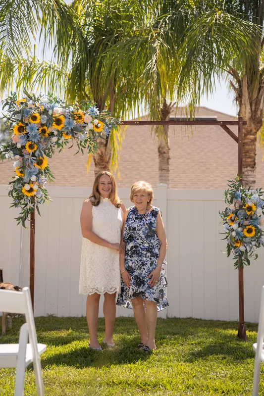 Two women stand together under the sunflower-adorned arch on the lawn, one in a white dress and one in a patterned blue dress.