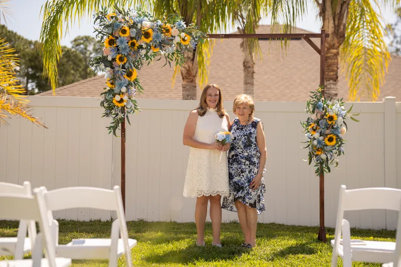 The two women stand hand-in-hand facing the camera beneath the flowering arch, with white ceremony chairs visible on either side.