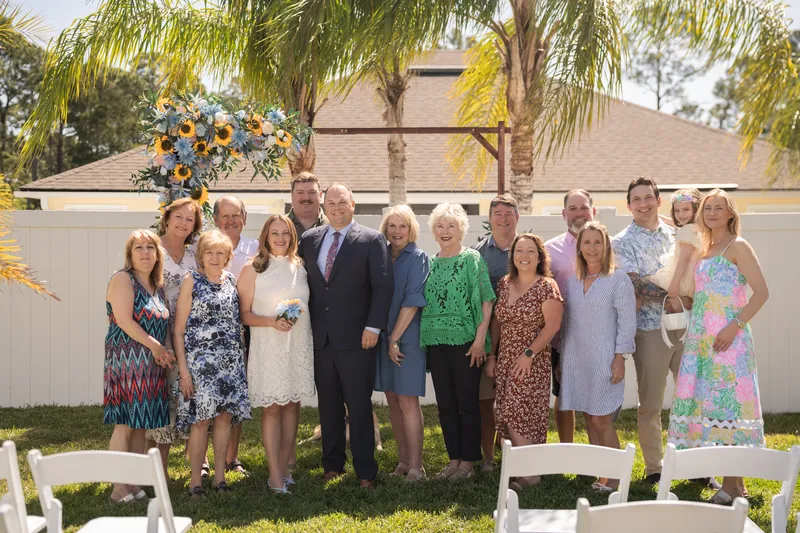 About 15 guests in bright summer clothes gather on a lawn beneath palm trees at the Airbnb in St. Augustine, with a sunflower arch behind them.