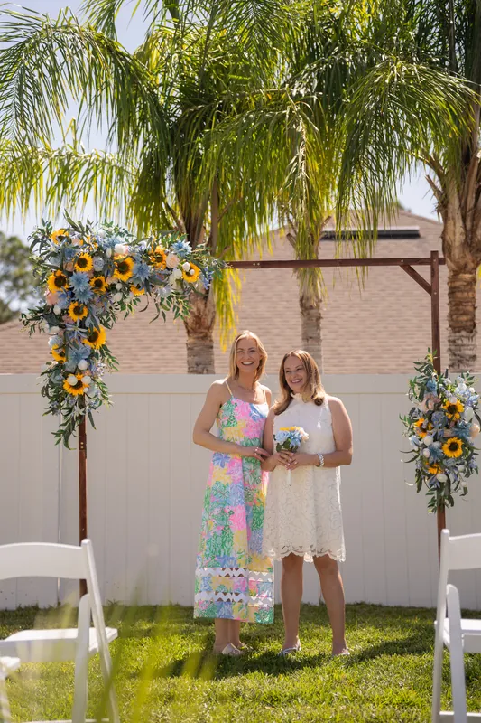 Two people in patterned dresses stand under a wooden arch decorated with sunflowers and greenery, framed by palm trees on a sunny lawn.