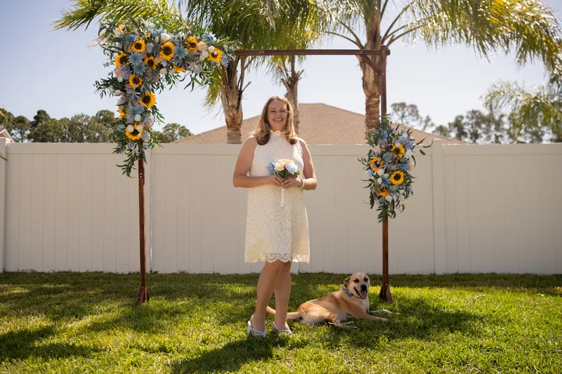 A person in a cream lace dress holds a white bouquet under a flower-draped wooden arch, with a brown dog sitting on the lawn beside them.