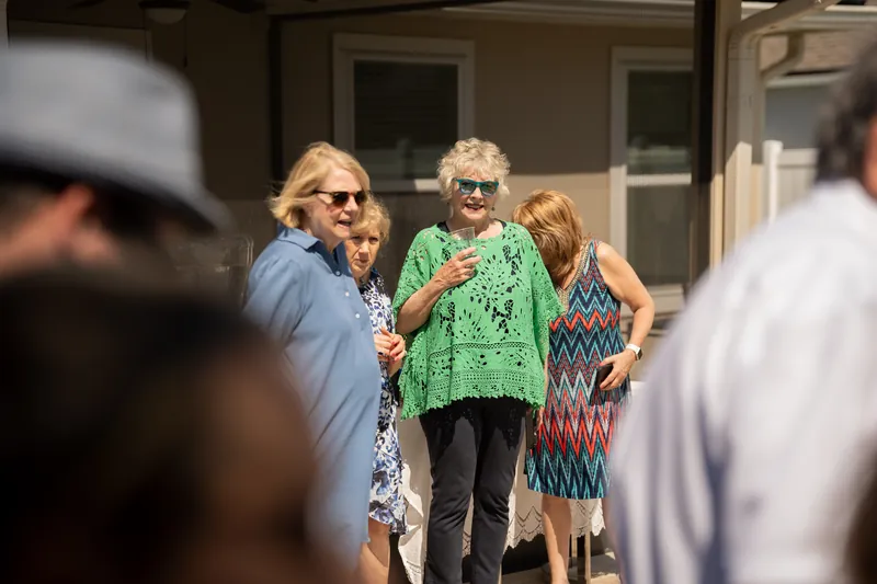 Several guests in summer clothing gather outdoors, some wearing sunglasses, with a house visible in the soft-focus background.