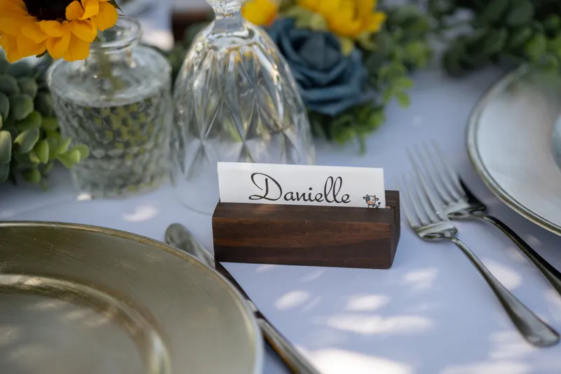 A place card reading "Danielle" in a wooden holder sits beside a plate setting with yellow and blue flowers in the table arrangement.