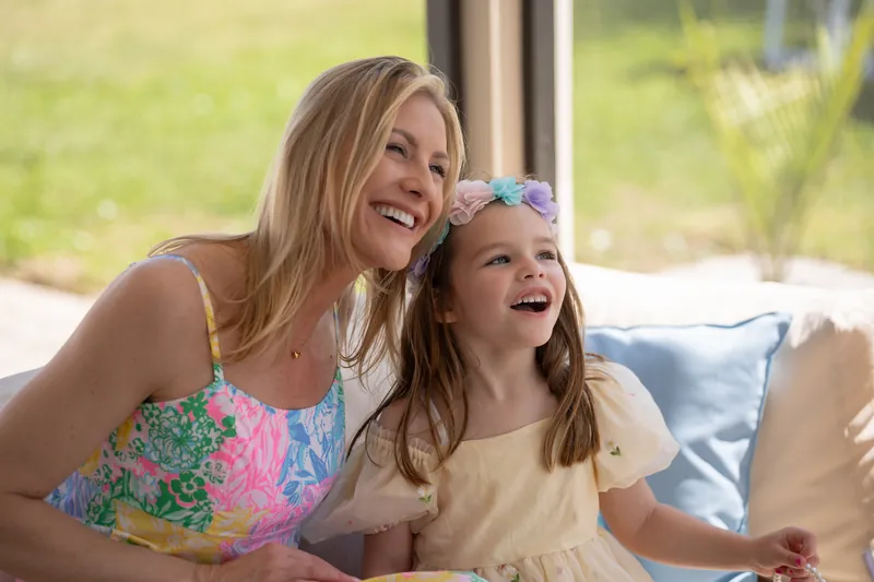 A woman in a colorful tie-dye dress laughs while sitting with a child wearing a blue headband indoors.