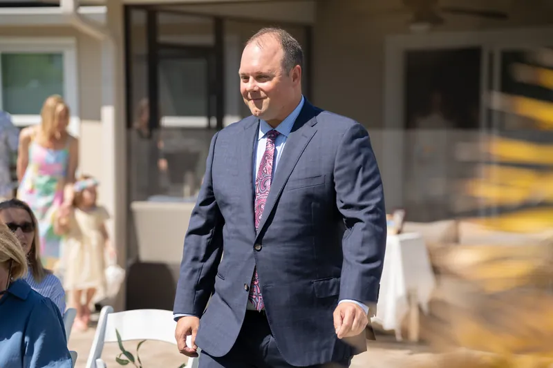 A person in a navy blazer and pink tie smiles while walking through a backyard reception area.