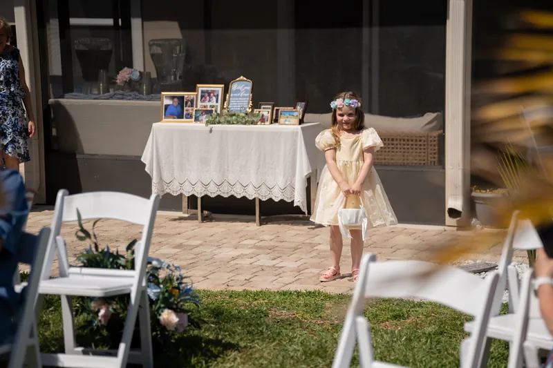 A child in a pale yellow dress stands near a white-clothed table displaying photos and flowers at an outdoor gathering.