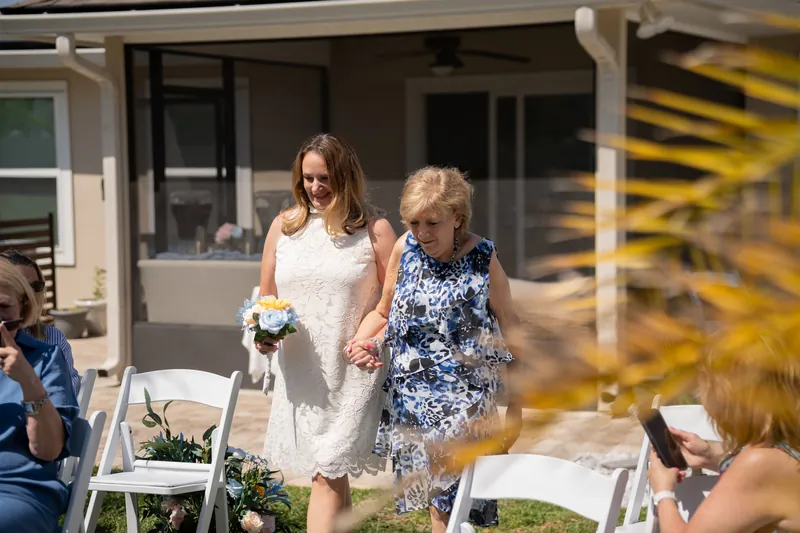 Two women in white dresses walk together holding a small bouquet during a backyard ceremony.