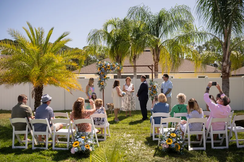 Guests seated in white chairs face one another across a grassy yard framed by palm trees under clear sky.