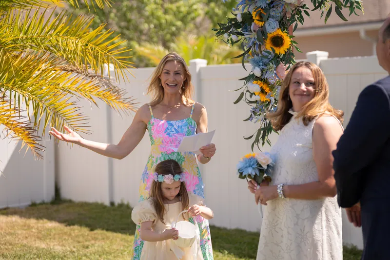 Two women and a child in a yellow dress stand together holding blue and yellow floral bouquets under palm fronds during the backyard ceremony.