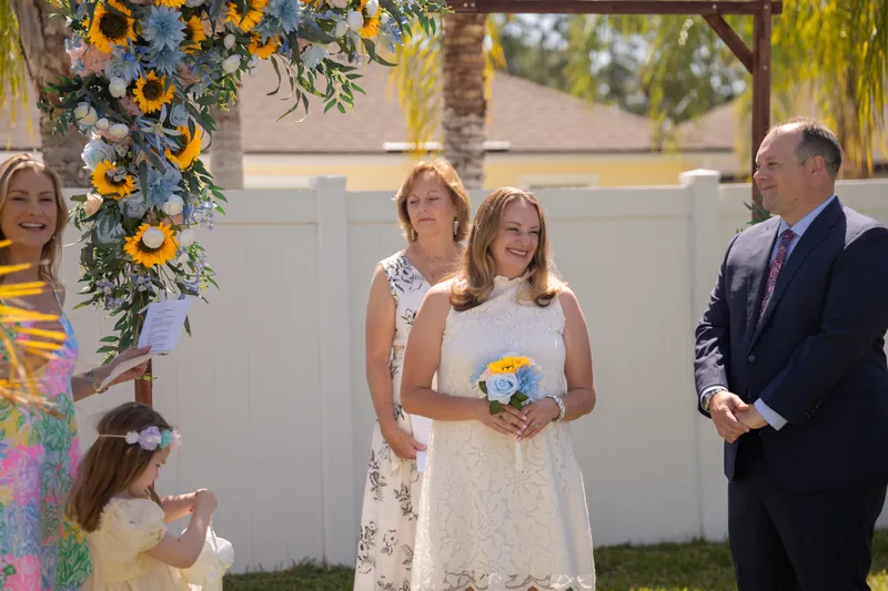 A child holds a small bouquet while standing next to two adults beside a colorful floral arch during the celebration.