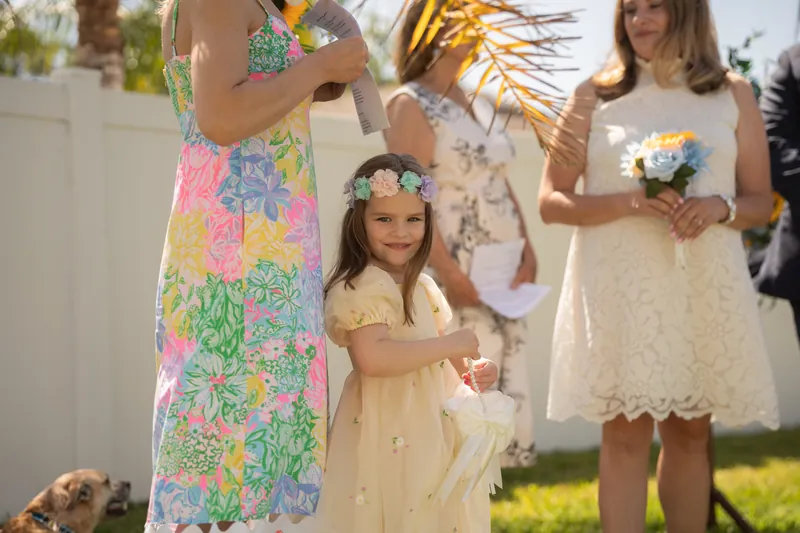 Guests in casual summer attire gather near the decorated arch, with a child in a pale yellow dress holding flowers among the group.