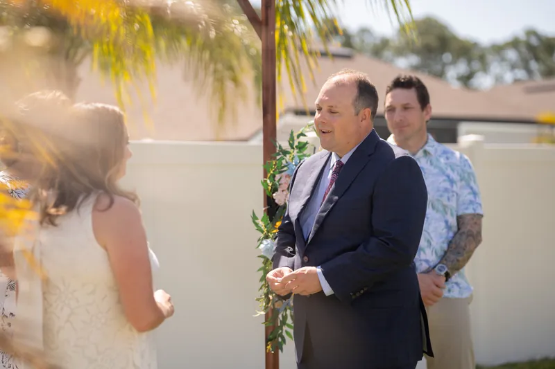 Two people exchange vows beneath a sunlit floral arch while a guest in a blue shirt stands nearby at the Airbnb in St. Augustine.