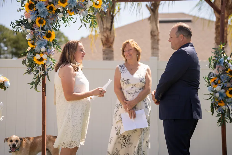 A woman in white listens as another woman speaks during the ceremony, with a man in navy and a small dog nearby under the flower-adorned arch.