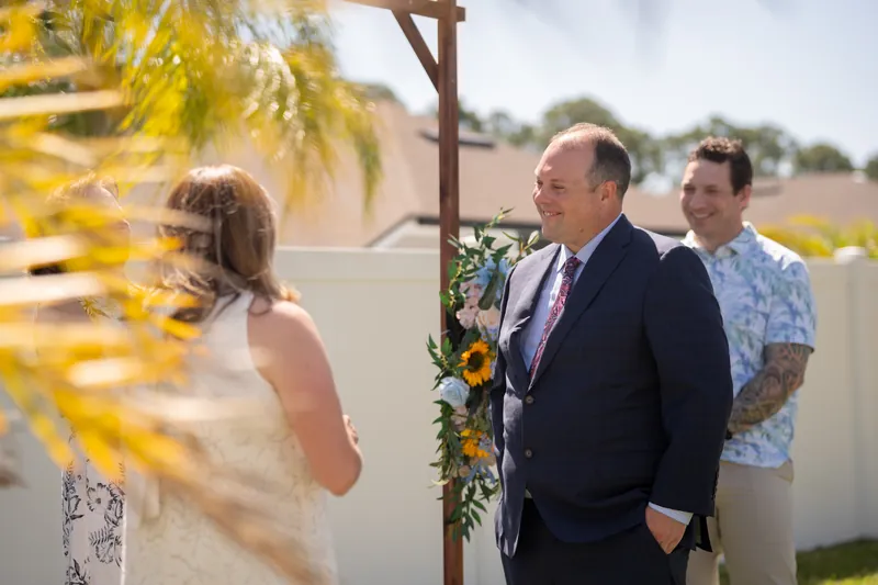 A person in a floral dress faces two others beneath the sunlit arch decorated with yellow and green blooms.